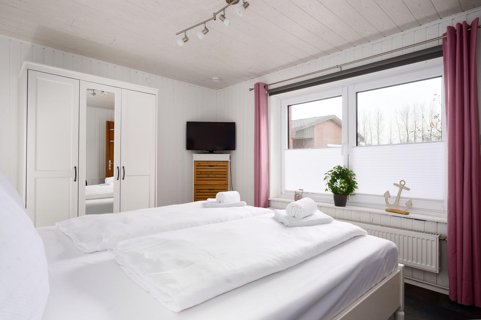 Schlafzimmer mit Doppelbett, weißen Wänden, TV und Fenster mit rosa Vorhängen.
