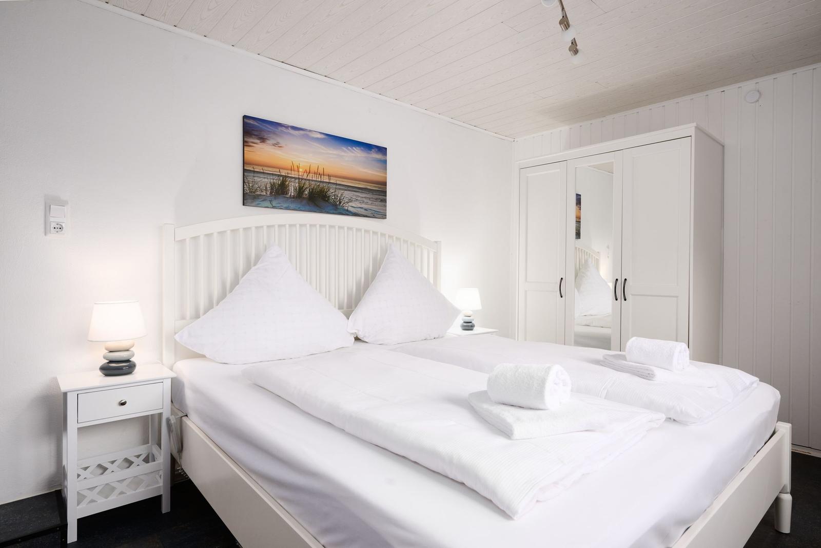 Schlafzimmer mit weißem Bett, Nachttisch und Kleiderschrank. Wandbild zeigt Strand.