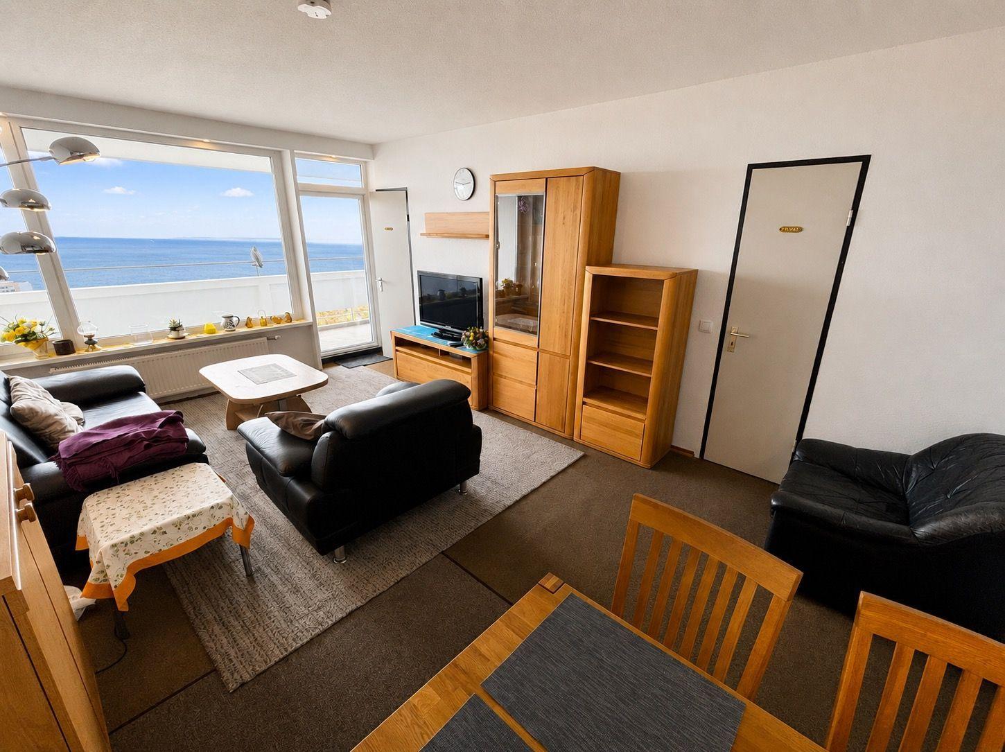 Wohnzimmer mit Blick auf Meer, schwarze Sitzgelegenheiten, Holzmöbel und Essbereich.