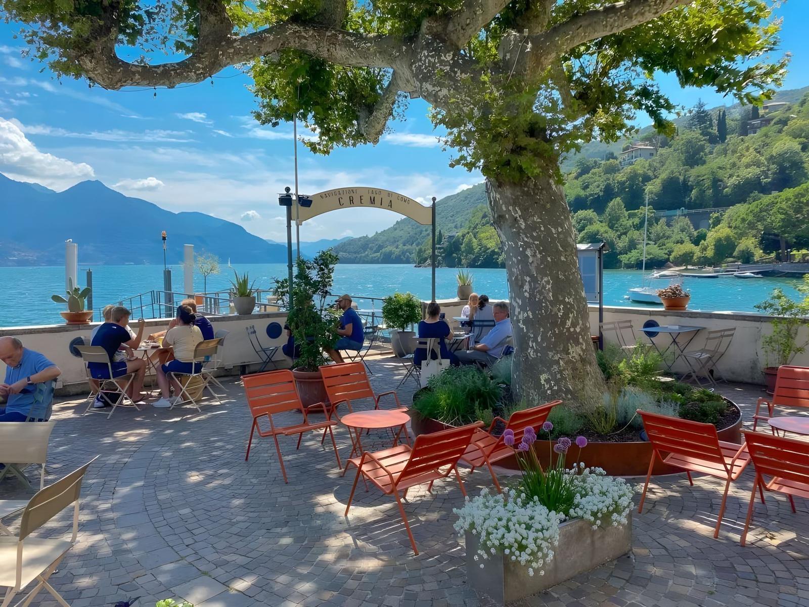 Terrasse mit orangefarbenen Stühlen und Blick auf den See. Großer Baum und Pflanzen sind sichtbar.