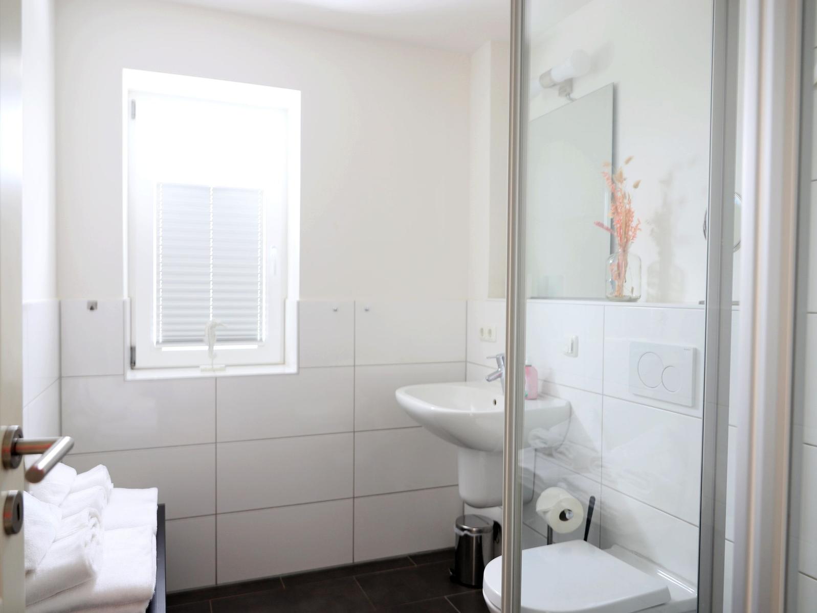 Badezimmer mit Dusche, Waschbecken und Fenster mit Jalousie.