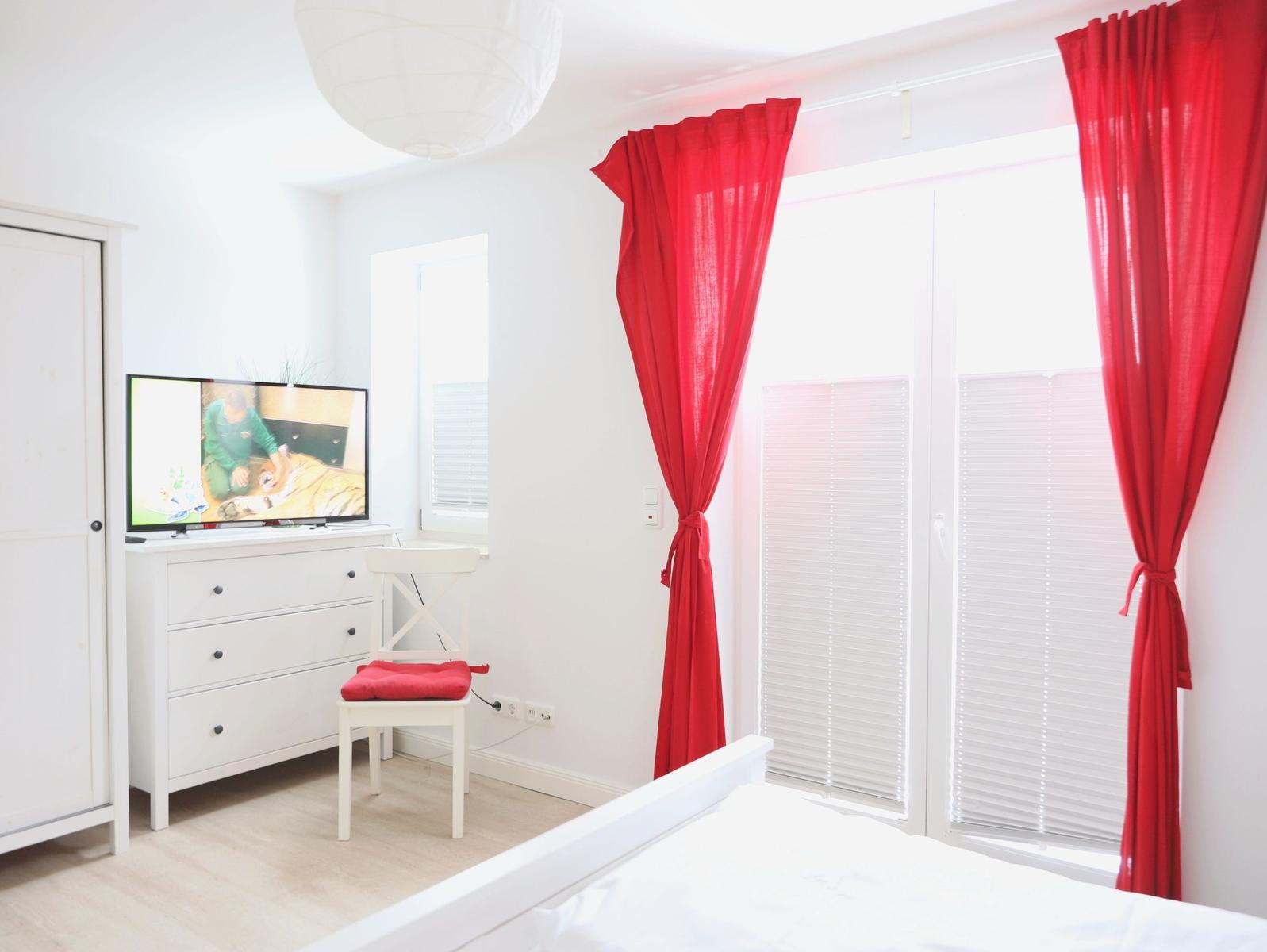 Zimmer mit weißem Bett, roten Vorhängen und Fernseher auf Schrank.