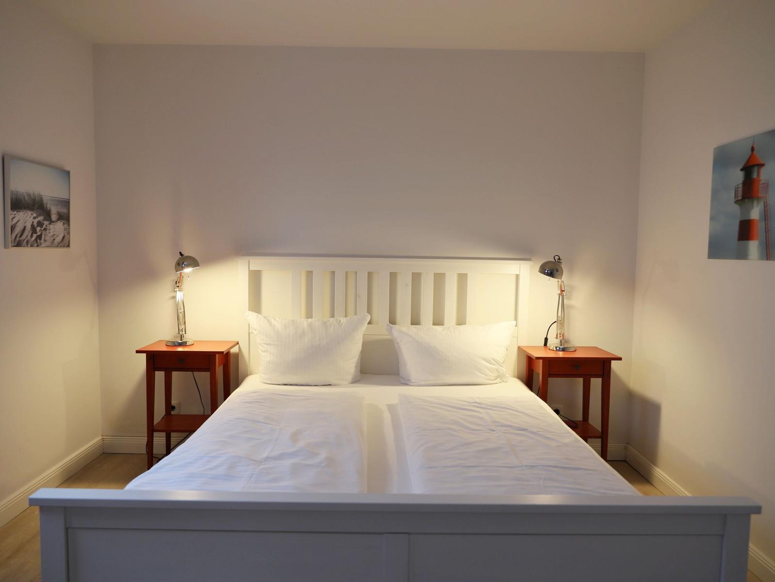 Schlafzimmer mit weißem Bett, zwei Nachttischen und Wandbildern.