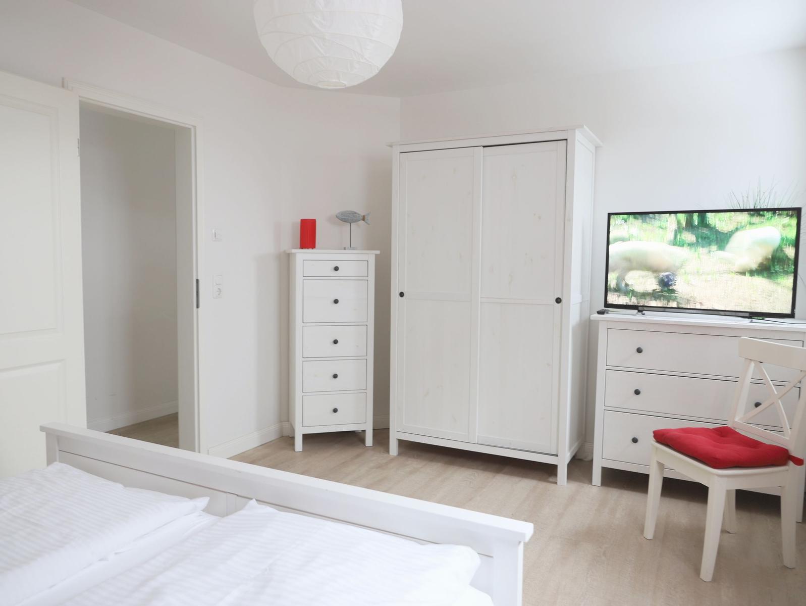 Schlafzimmer mit weißem Bett, Kleiderschrank, TV und Stuhl mit rotem Sitzkissen.