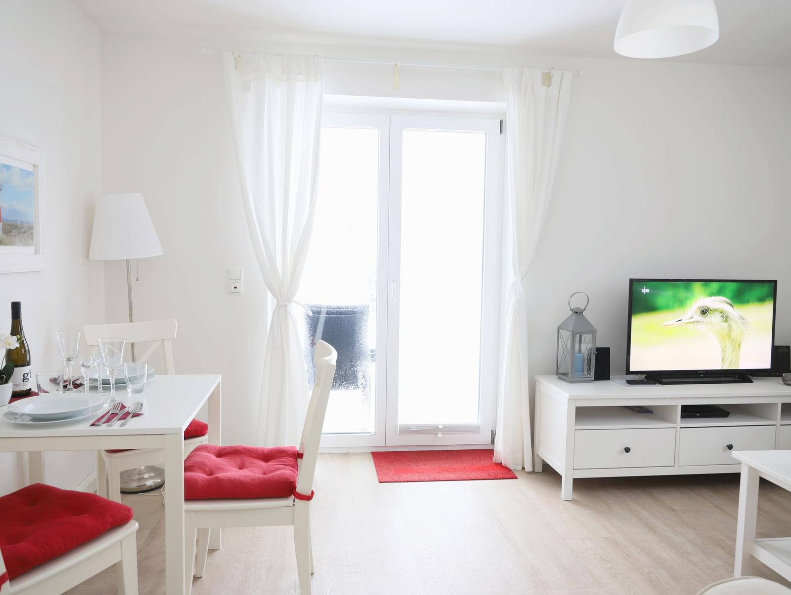 Wohn- und Essbereich mit TV, Tisch, Stühlen und Fenster. Rote Sitzkissen und Teppich.