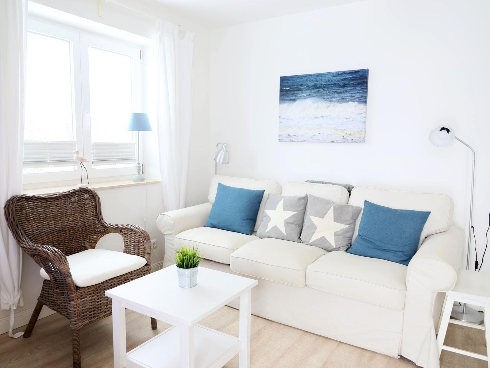 Wohnzimmer mit weißem Sofa, blauen und grauen Kissen, Wickerstuhl und Wandbild mit Wellen.