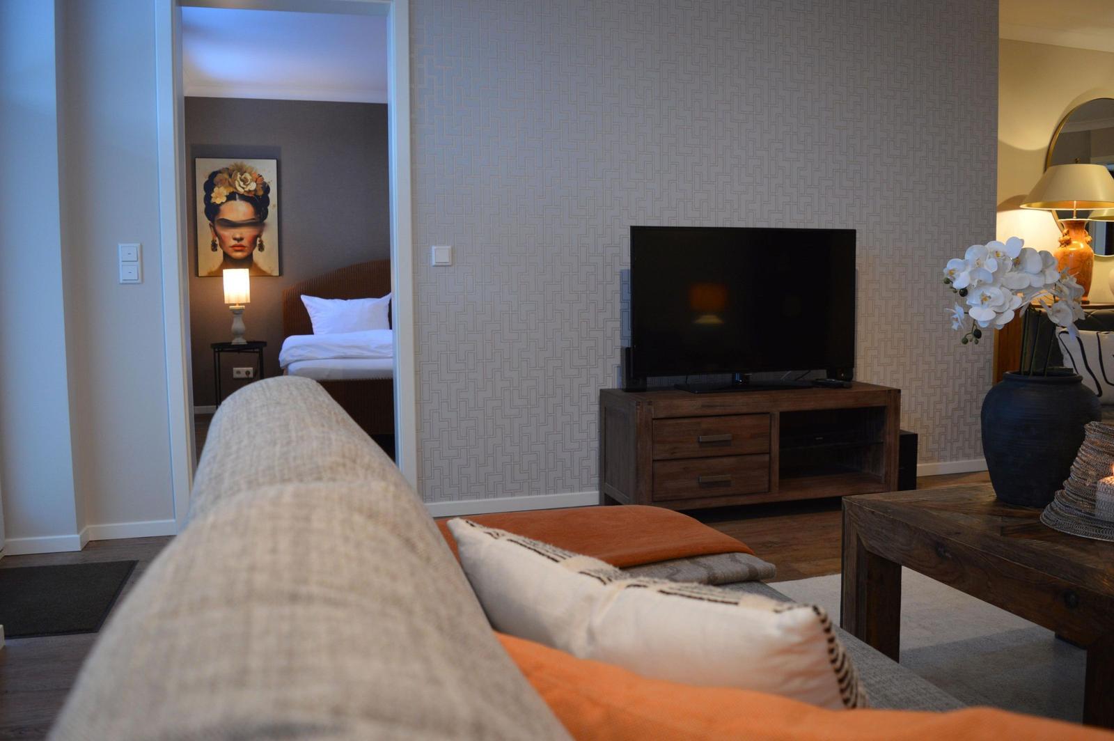 Wohnzimmer mit Sofa, Fernseher und Blick in Schlafzimmer mit Bett und Wandbild.