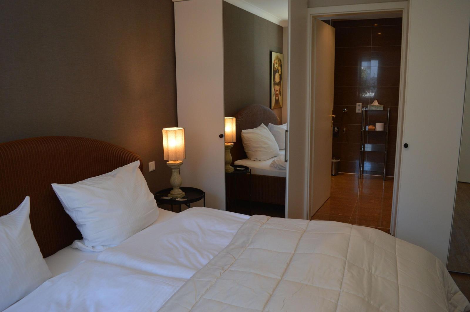 Schlafzimmer mit Bett, Nachttisch und Lampe. Durchgang zur Dusche mit dunkler Fliesenwand.