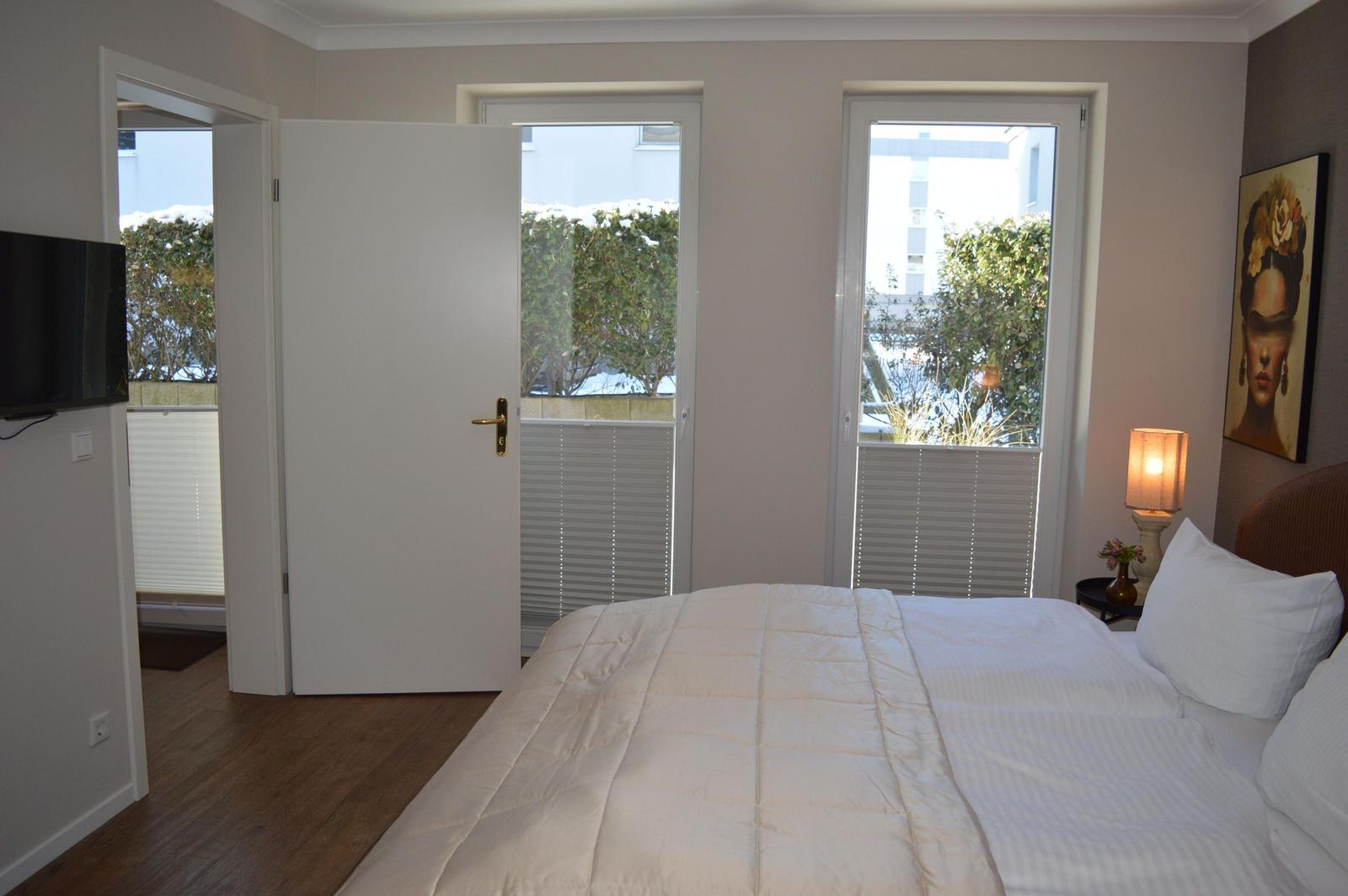 Schlafzimmer mit Bett, Fenster und Tür. Wandbild und Nachttisch mit Lampe.
