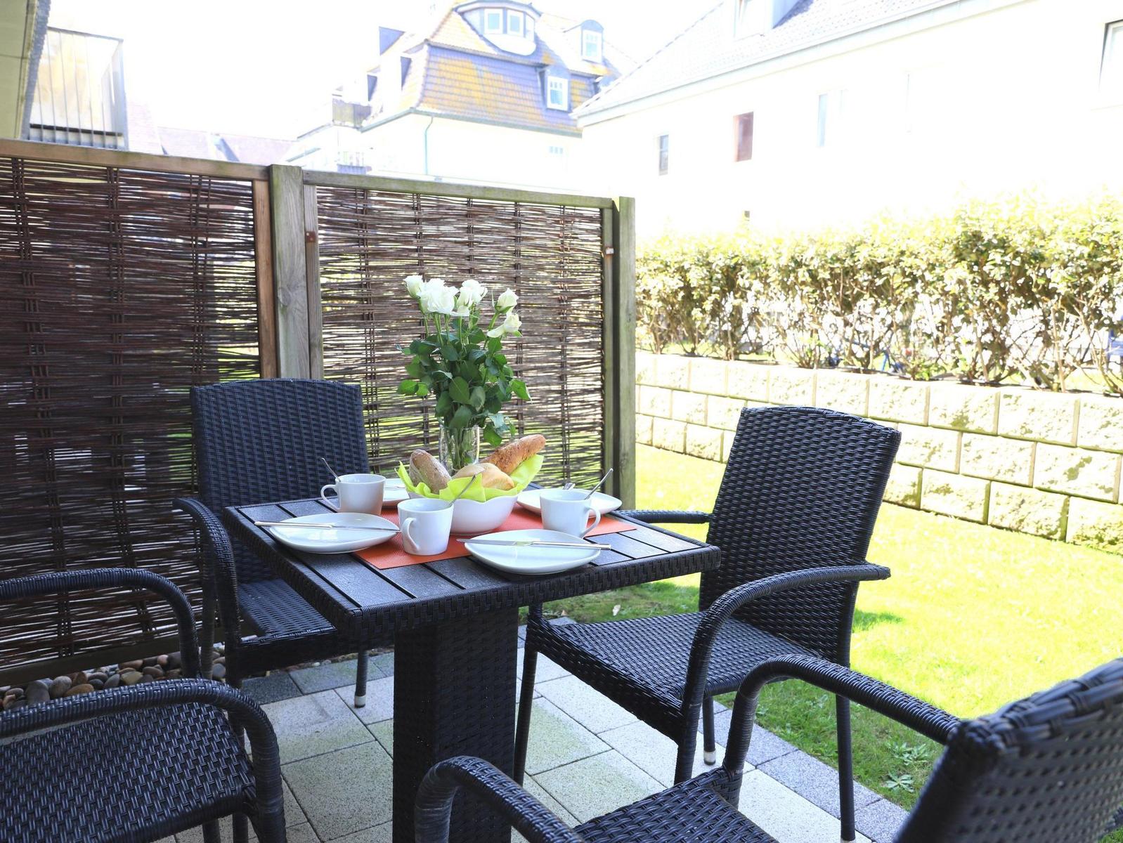 Terrasse mit Tisch, Stühlen und Frühstückszubehör. Hintergrund: Garten und Gebäude.
