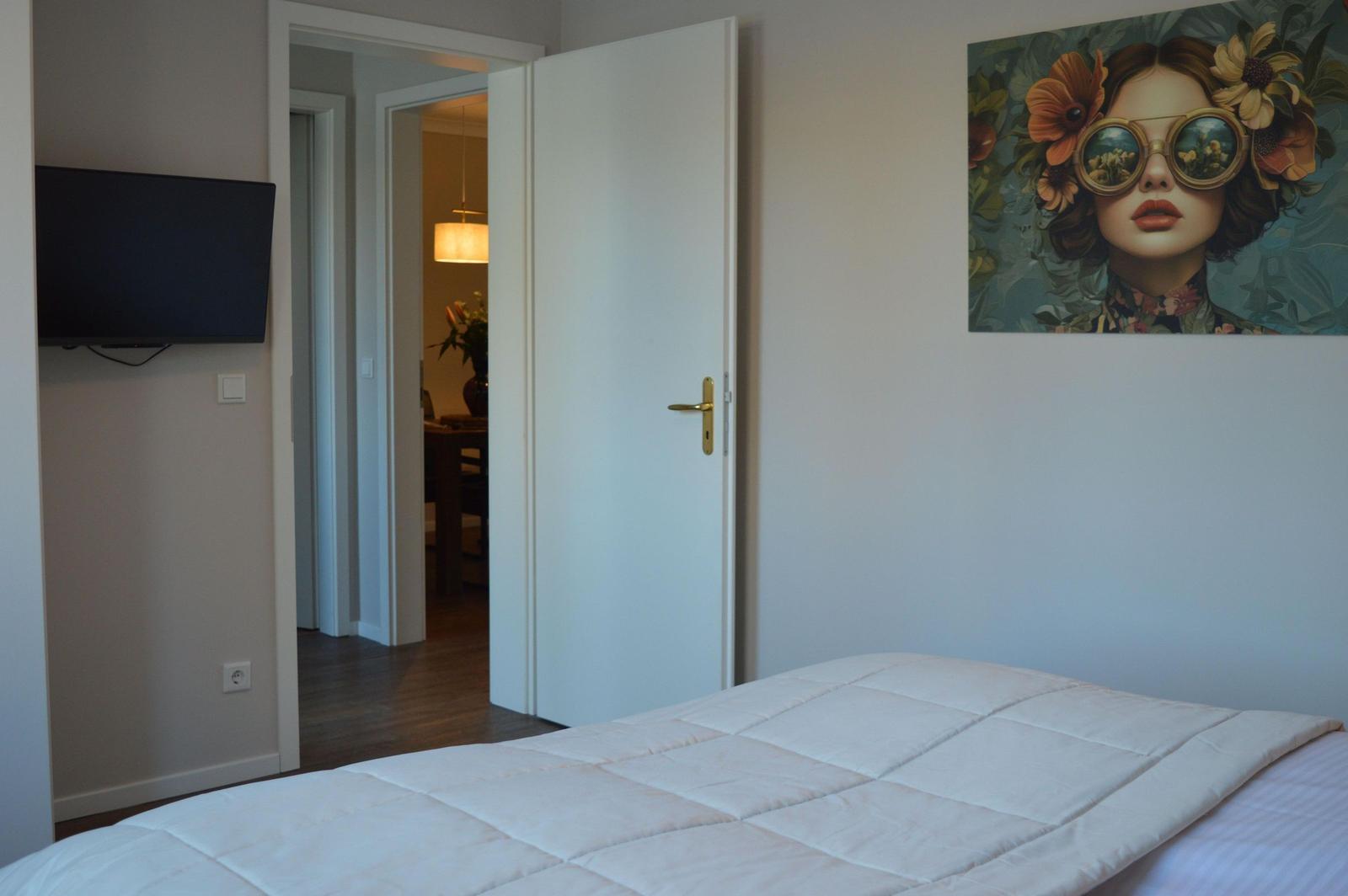 Schlafzimmer mit Bett, Wandbild und Fernseher. Tür führt in angrenzenden Raum.