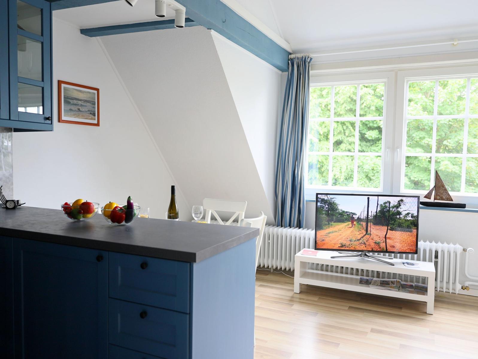 Küche mit blauen Schränken und Essbereich mit TV und Fenster.