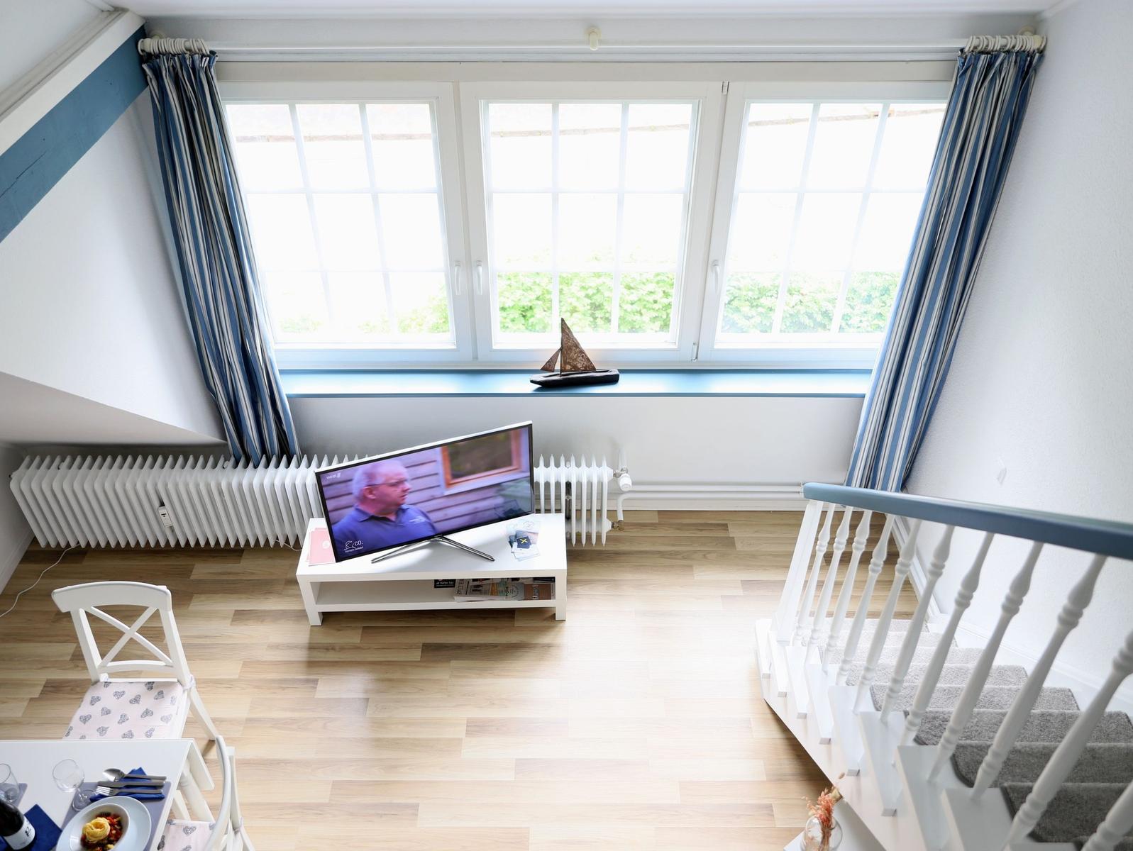 Wohnraum mit TV, Fenster und Treppe. Holzboden und weiße Möbel.