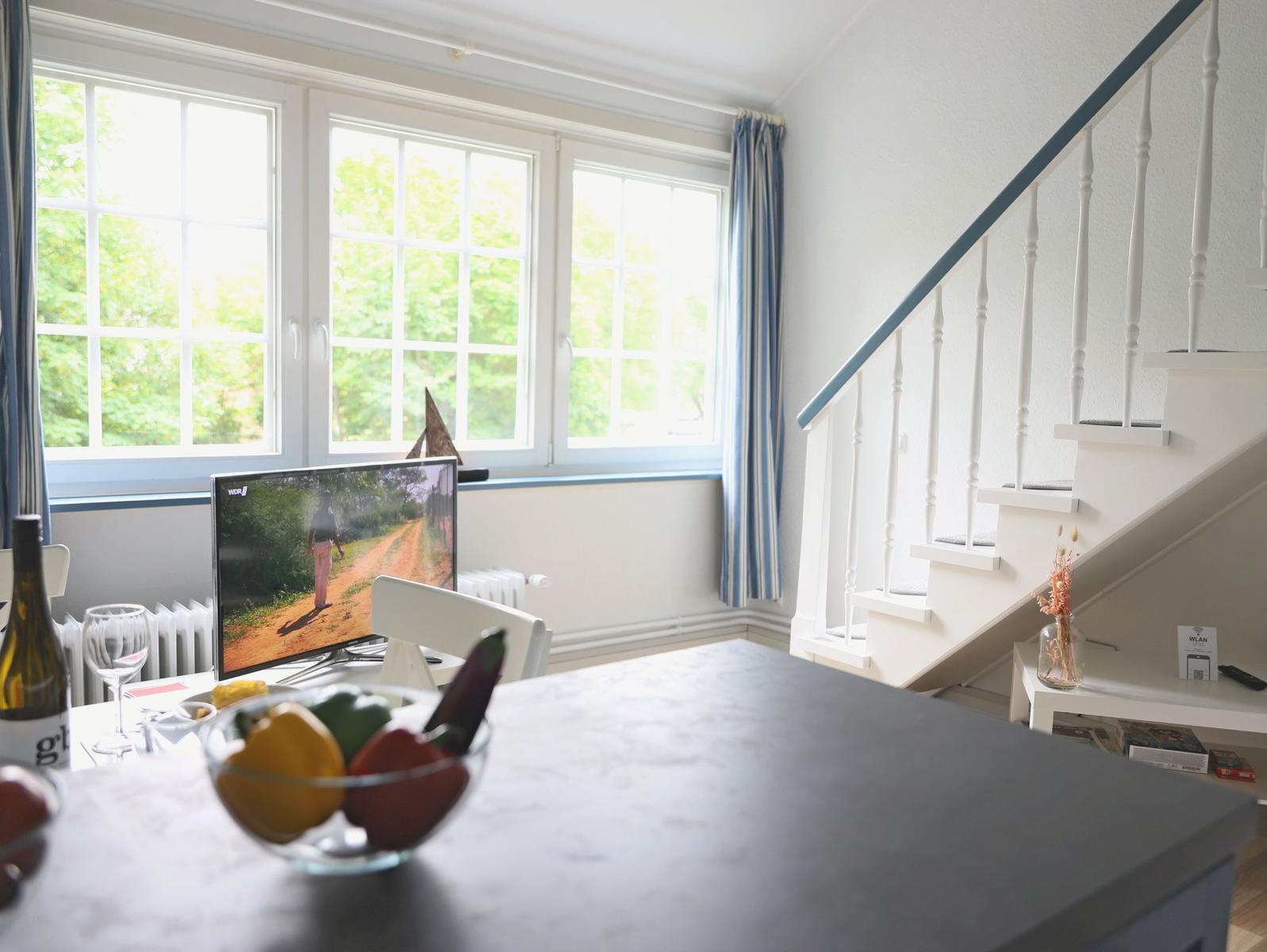 Ein Essbereich mit Tisch, Fernseher und Treppenhaus. Fenster mit Blick auf Bäume.