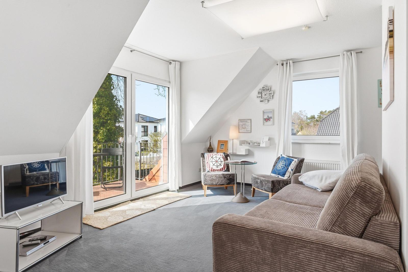 Wohnzimmer mit Sofa, Sesseln und TV. Große Fenster und Balkontür. Dachschrägen.