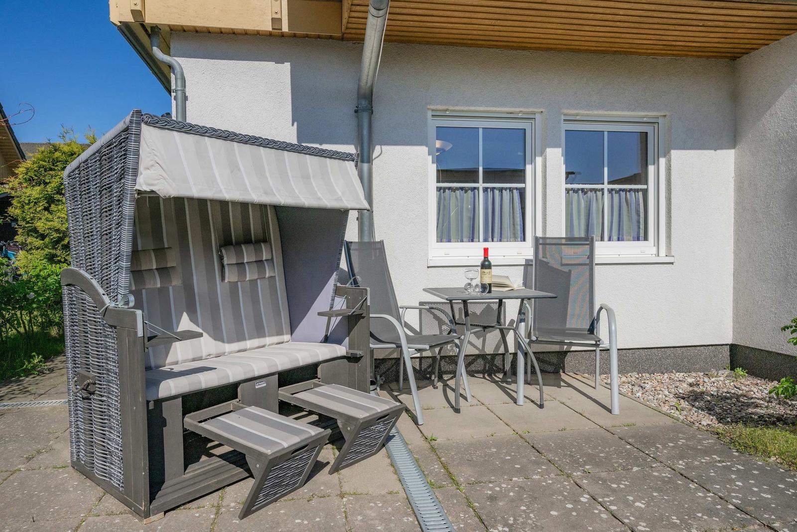 Terrasse mit Strandkorb, Tisch und zwei Stühlen. Hintergrund: Haus mit Fenstern.