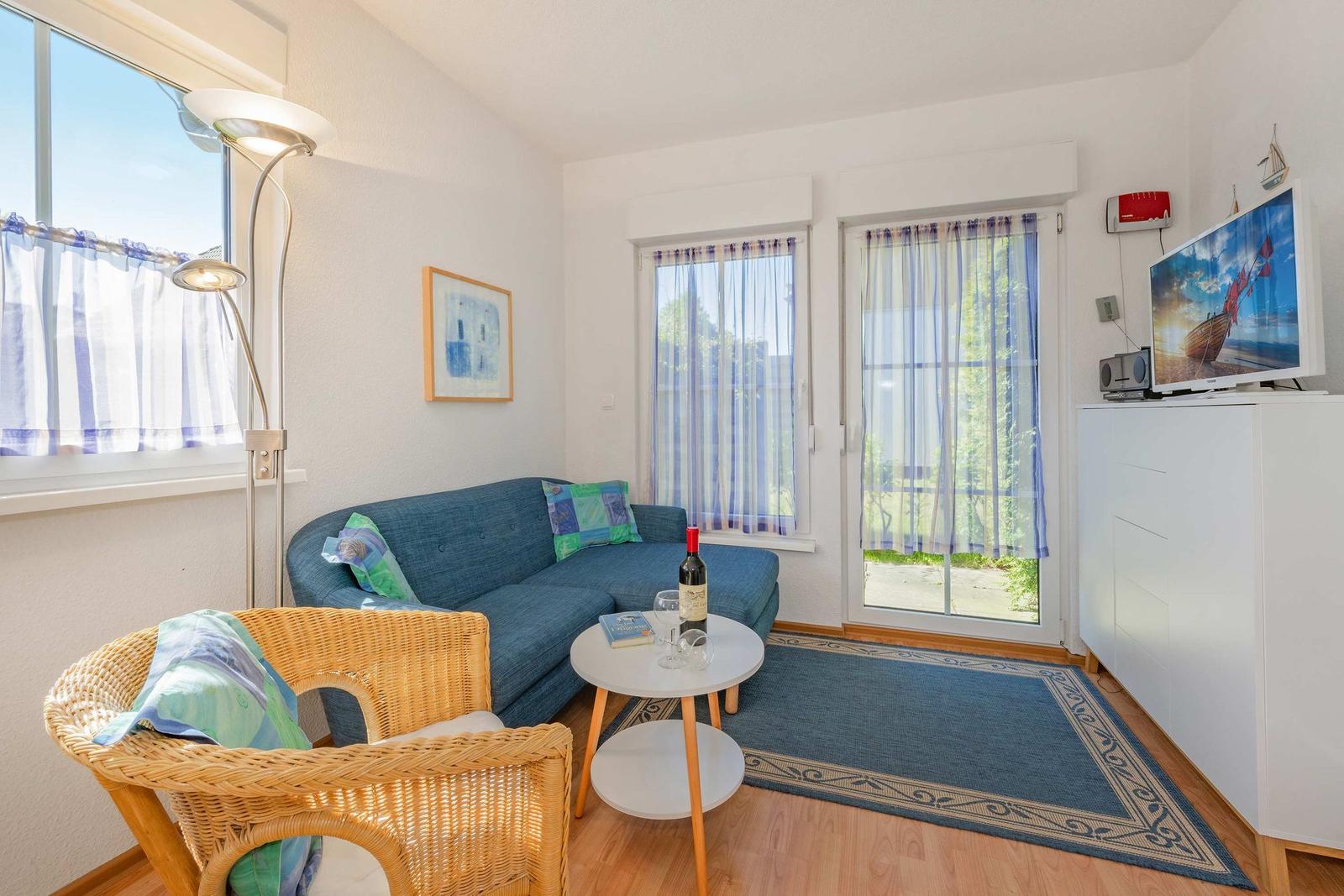 Ein Wohnzimmer mit Sofa, Stuhl, Tisch und Fernseher. Fenster und Tür bieten Blick auf Außenbereich.