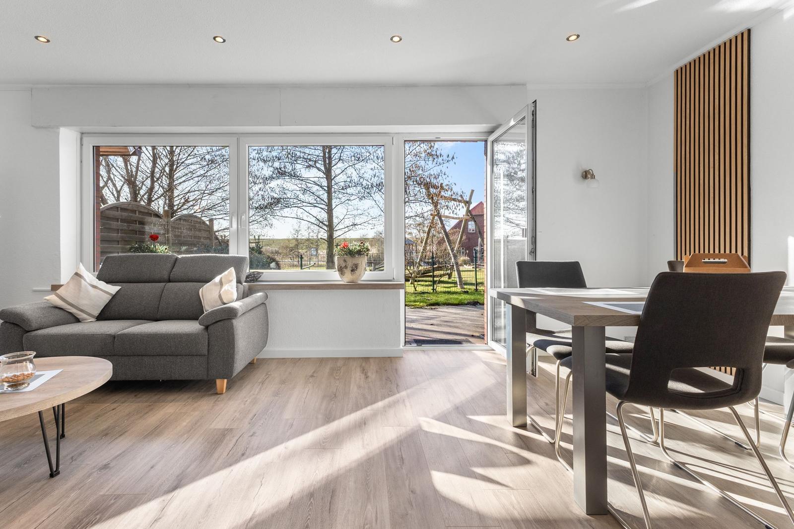 Wohn- und Essbereich mit Sofa, Tisch und Stühlen. Große Fenster und Tür zur Außenterrasse.