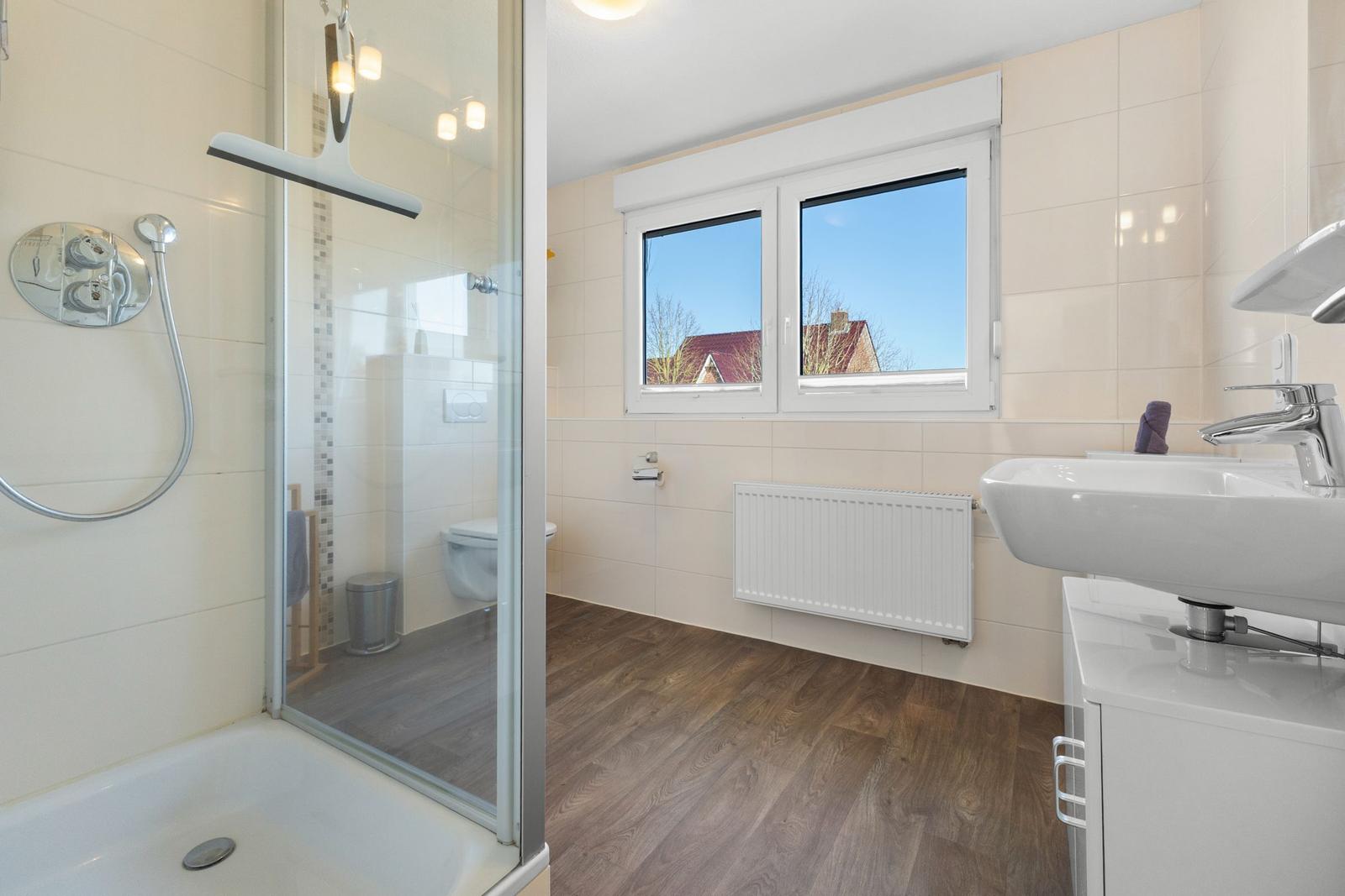 Badezimmer mit Dusche, Waschbecken und Fenster mit Blick auf Dächer.