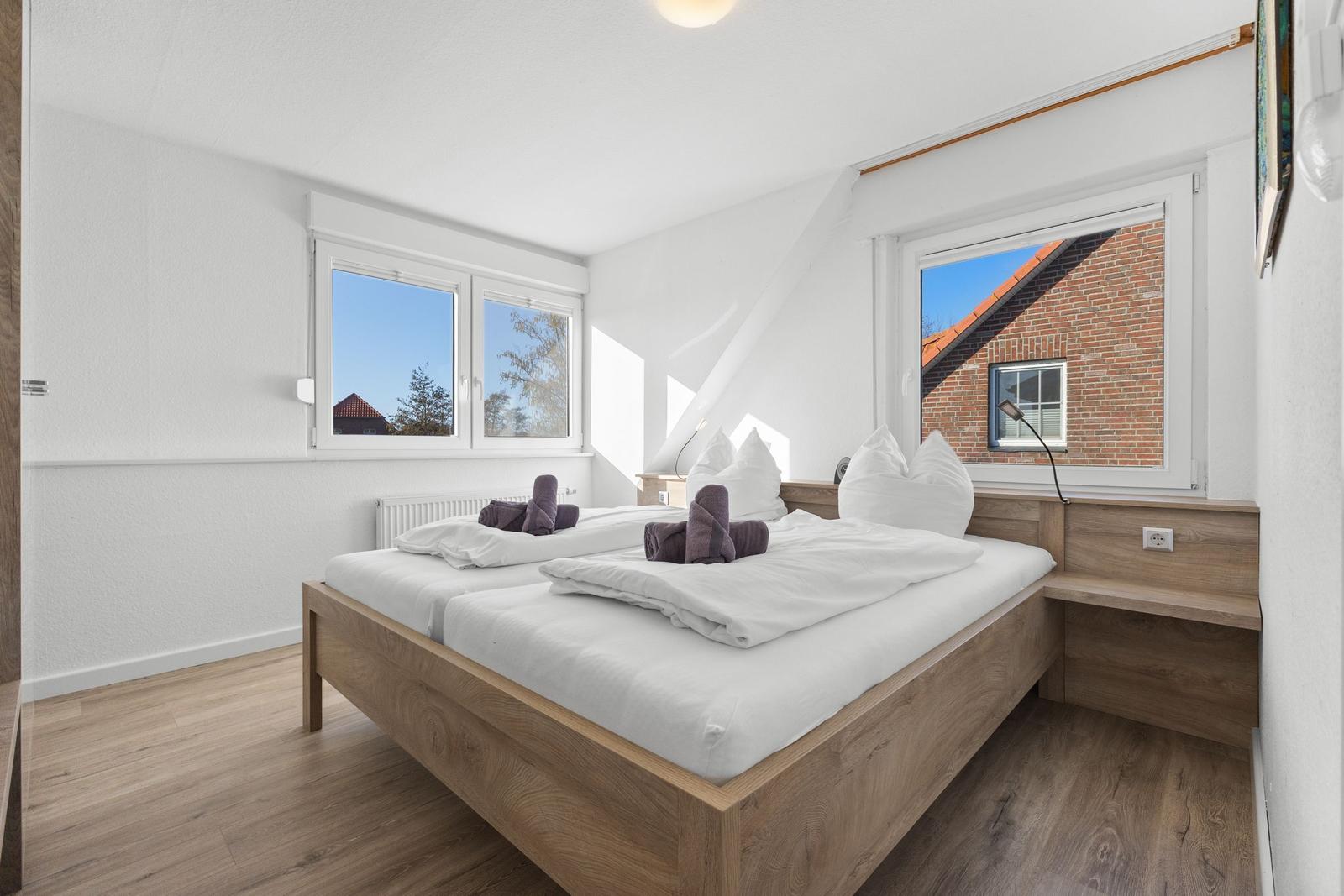Doppelbett mit weißen Laken und Lakenrollen. Zwei Fenster mit Blick auf Dächer.