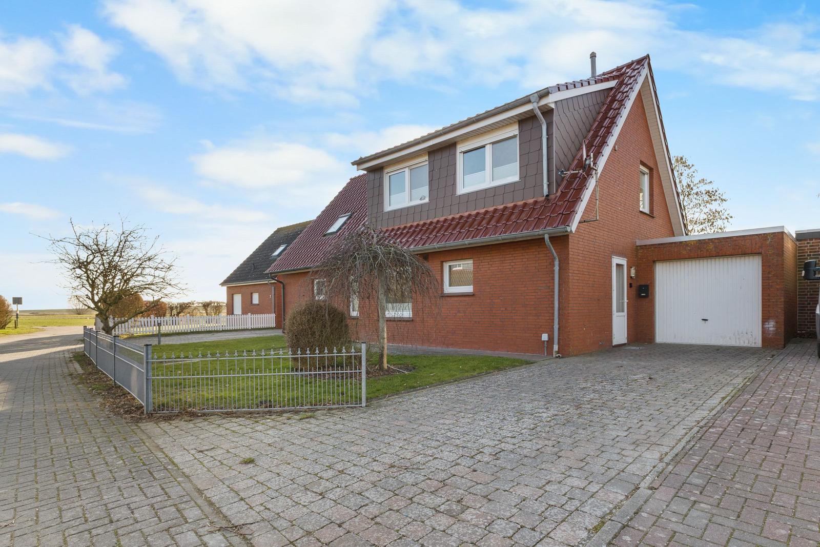 Zweistöckiges Haus mit Garage und gepflastertem Vorplatz. Umgeben von Grünfläche und Zaun.