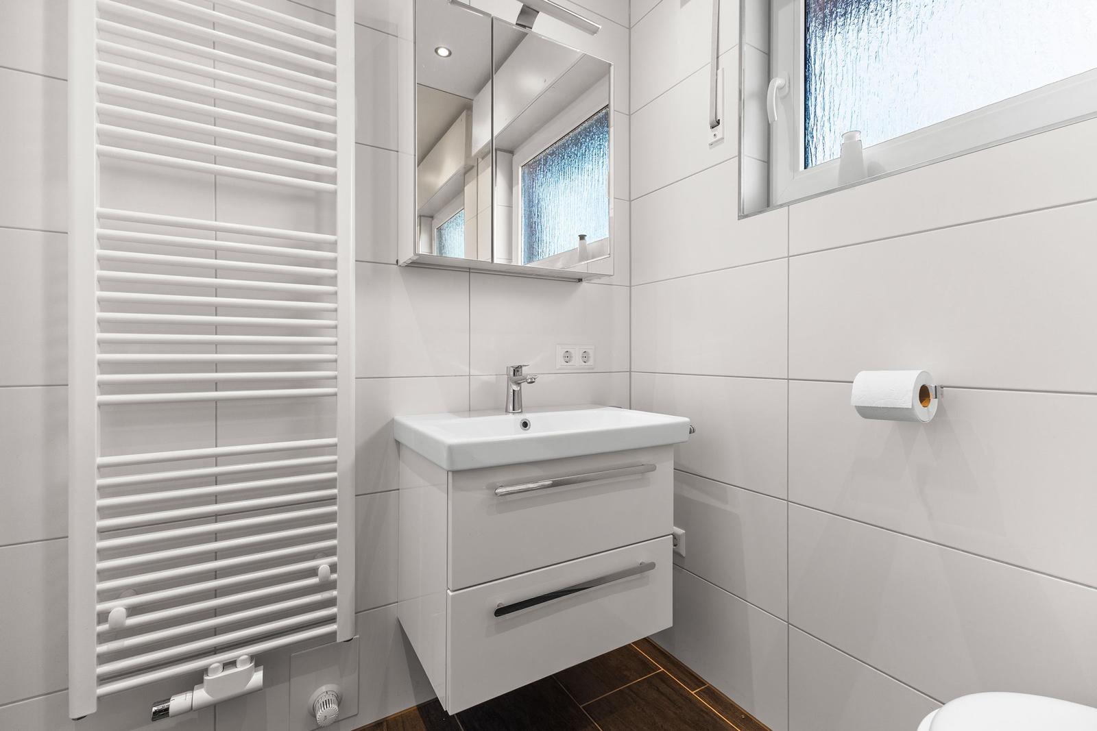 Badezimmer mit Waschbecken, Spiegel, Heizkörper und Toilettenpapierhalter.
