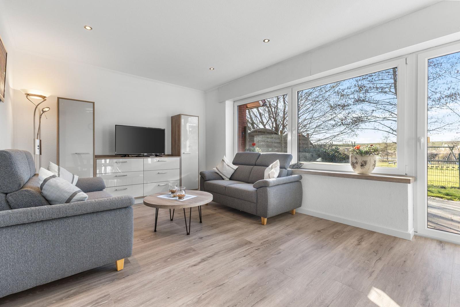 Wohnzimmer mit grauen Sofas, Holzboden und großem Fenster zur Außenterrasse.