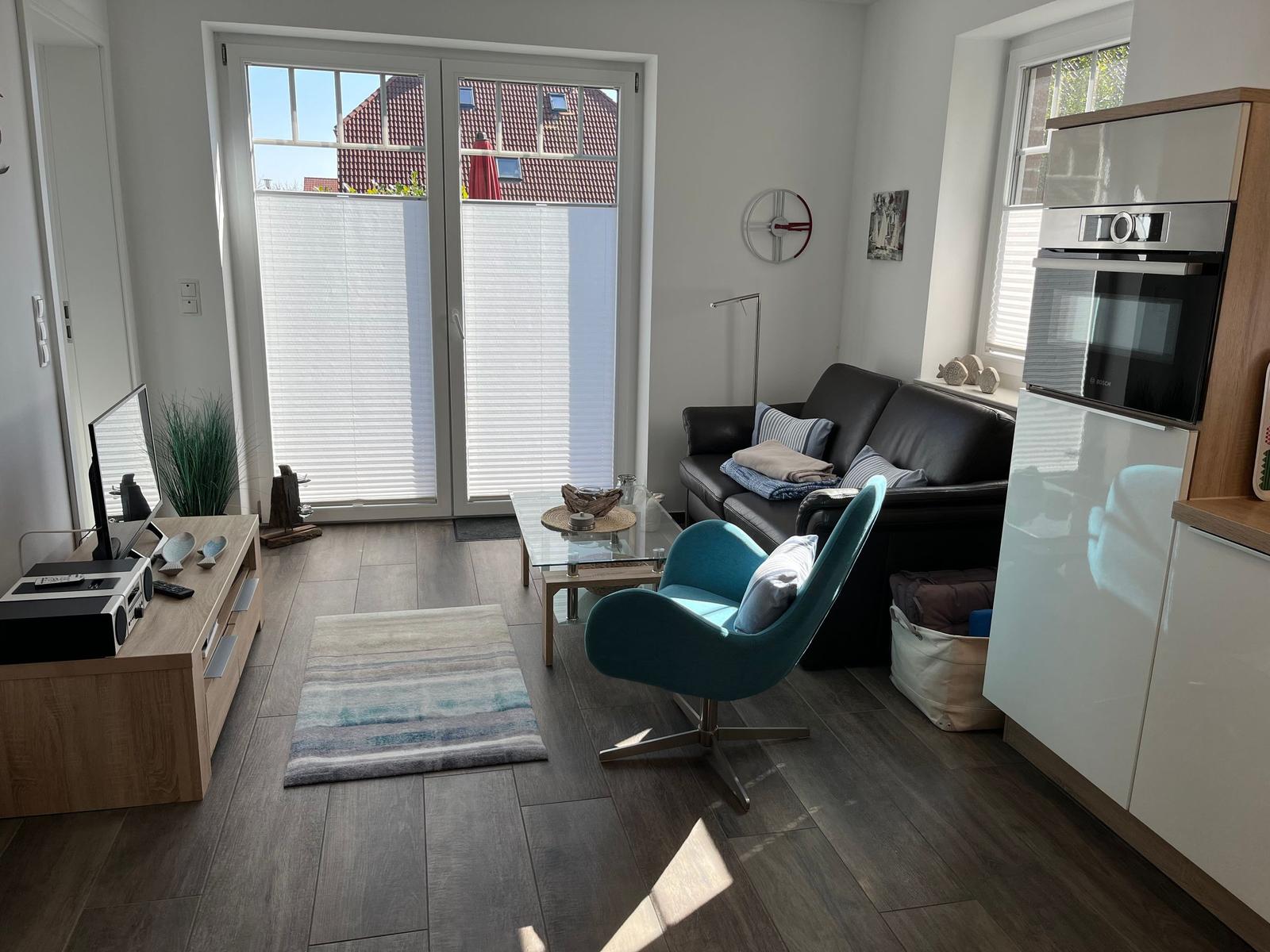 Wohnzimmer mit Sofa, Sessel und Küchenzeile. Durchgang zur Terrasse mit Fenster.