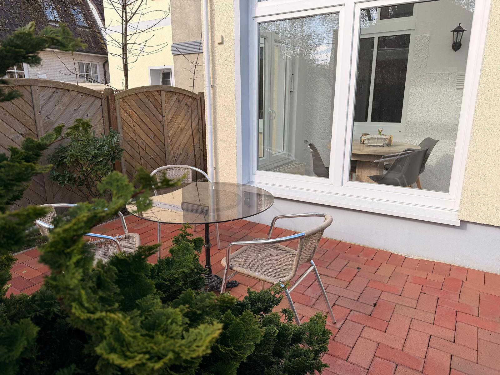 Terrasse mit Glas-Tisch und Stühlen, rote Pflastersteine und Holzzaun im Hintergrund.
