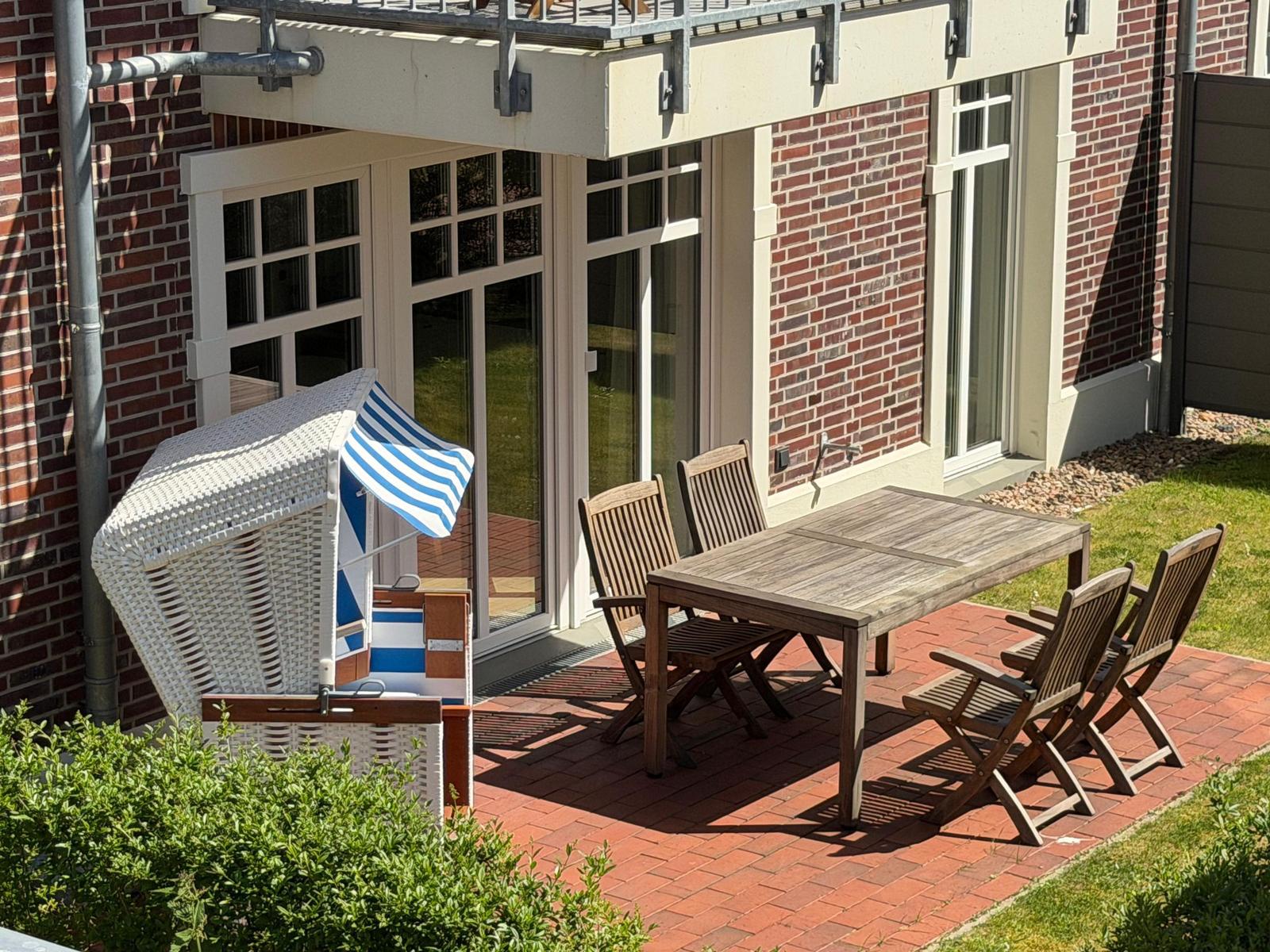 Terrasse mit Holztisch, vier Stühlen und Strandkorb. Hintergrund: rote Ziegelwand und Glasfront.