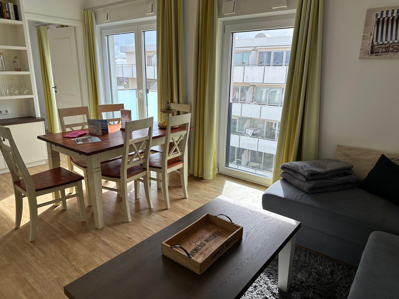 Wohn- und Essbereich mit Tisch, Stühlen und Sofa. Große Fenster mit Blick auf Gebäude.