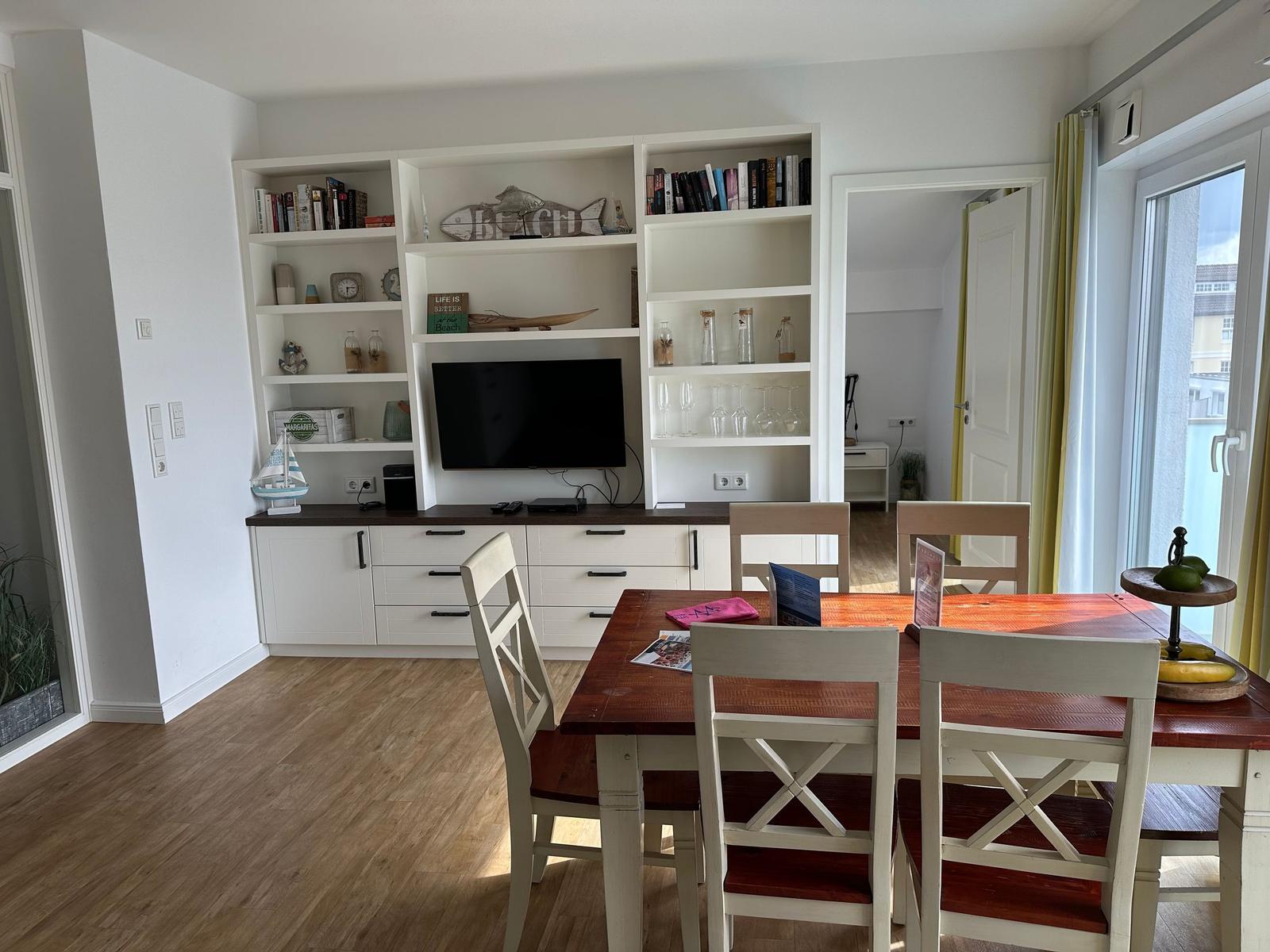Essbereich mit Tisch und Stühlen. Hintergrund: Bücherregal mit Fernseher und Glasflaschen.