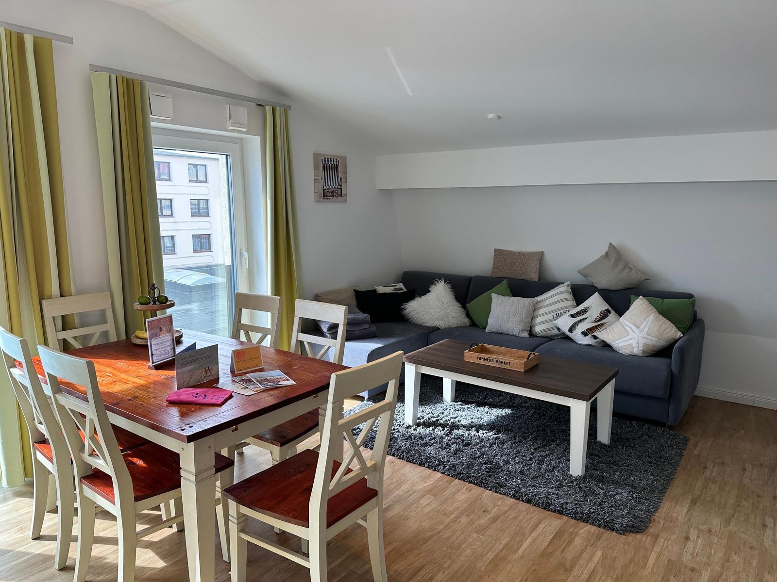 Wohn- und Essbereich mit Sofa, Tisch und Stühlen. Fenster mit grünen Vorhängen.