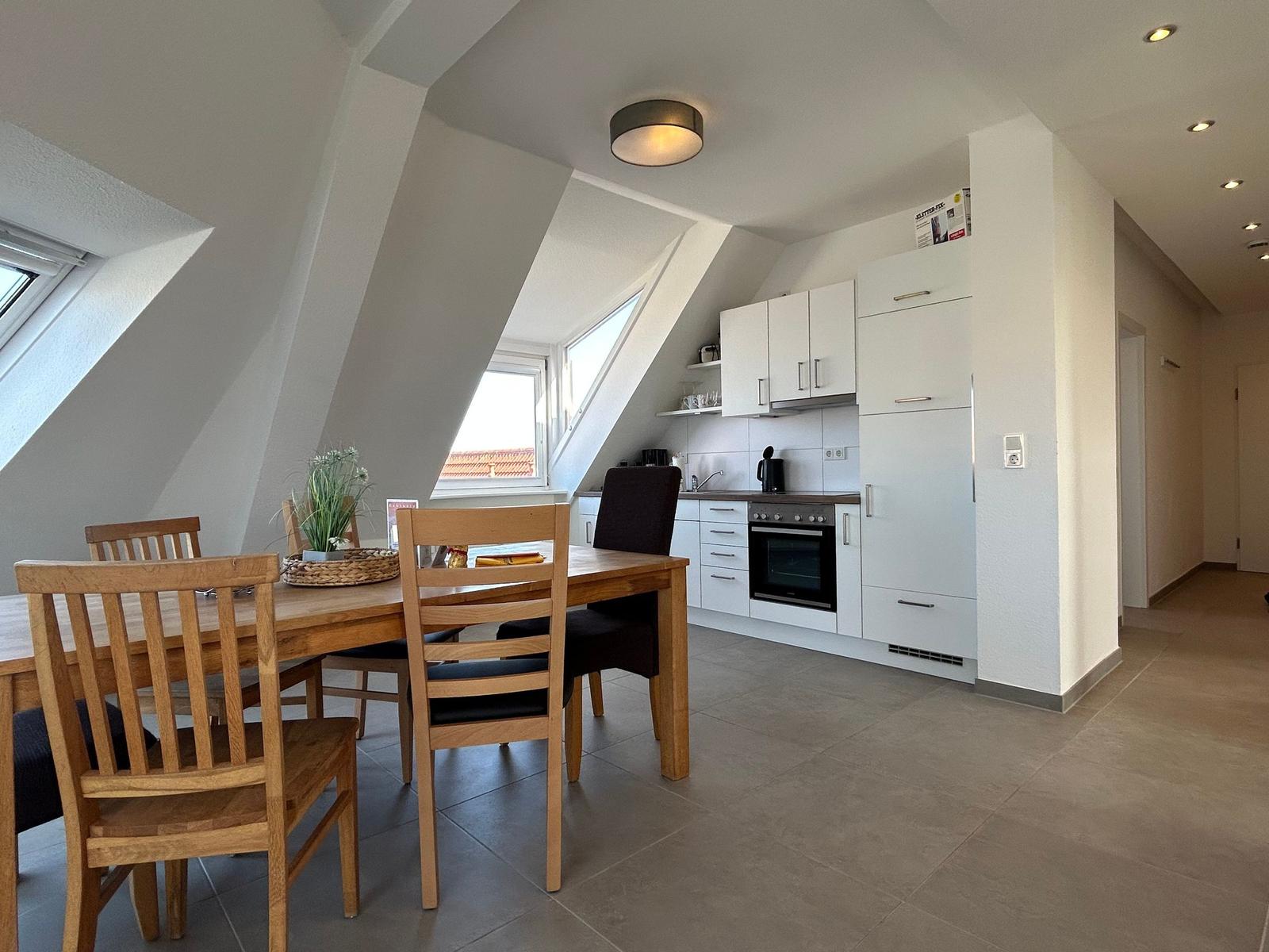 Essbereich mit Holztisch und Stühlen, angrenzende Küche mit weißen Schränken und Fenster.