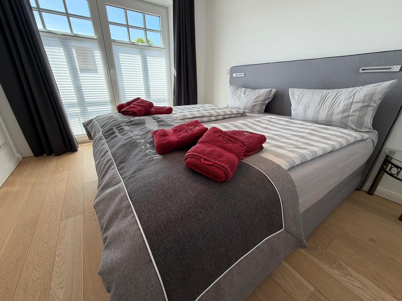 Schlafzimmer mit Doppelbett, grauem Kopfteil und roten Handtüchern auf der Bettdecke.