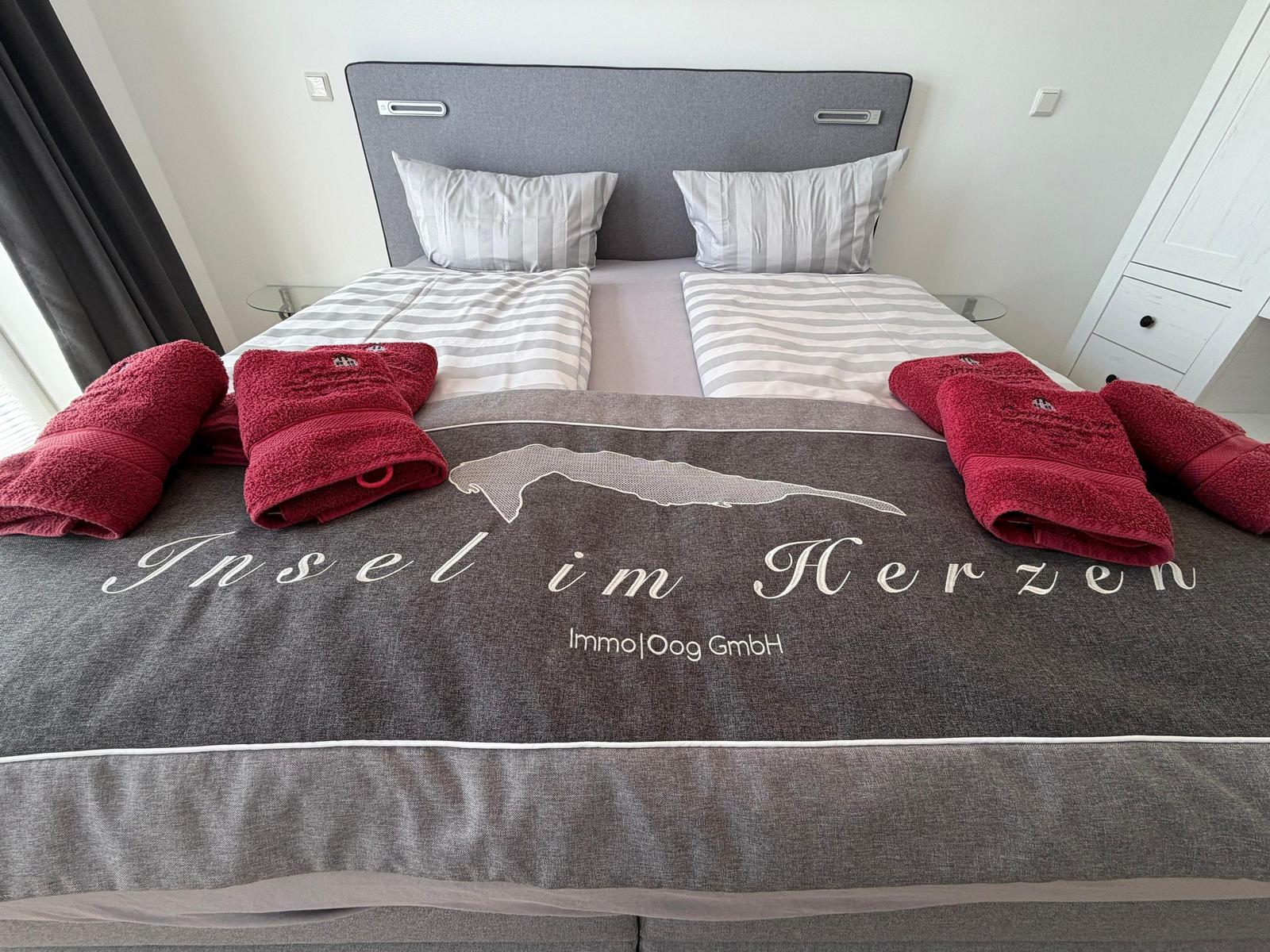 Doppelbett mit grauer Bettwäsche und roten Handtüchern. Kopfteil und Nachttische sichtbar.