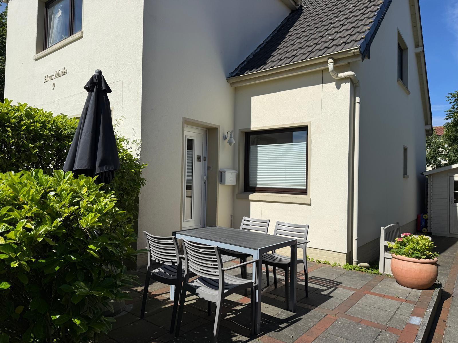 Haus mit Terrasse, Tisch, Stühlen und Schirm. Pflanzen und gepflastertes Gelände.