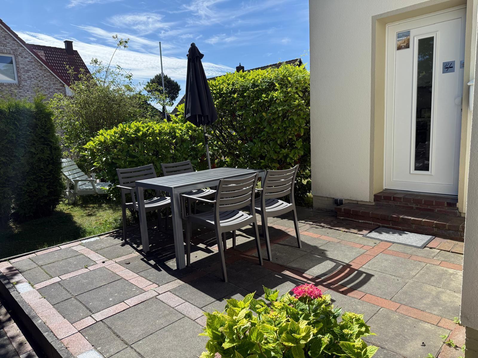 Terrasse mit Tisch, vier Stühlen und geschlossenem Sonnenschirm. Hinterhof mit Beete und Gartenhaus.