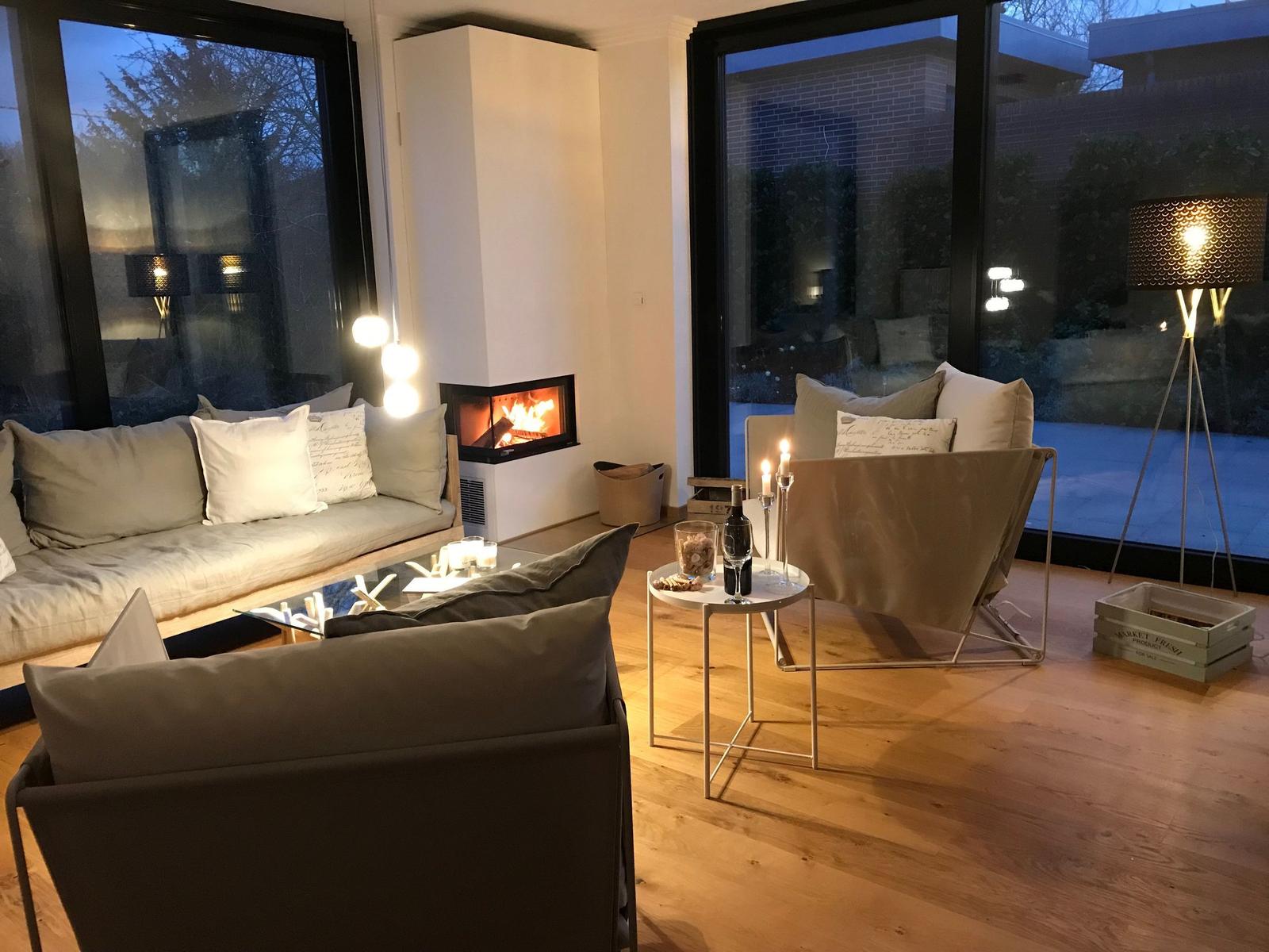 Wohnzimmer mit Kamin, Sofa und Sessel. Holzboden und große Fenster.