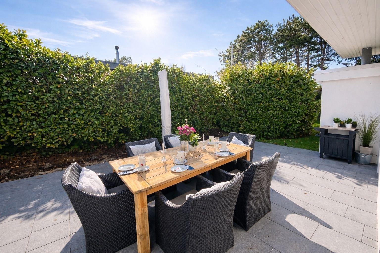 Terrasse mit Holztisch und Stühlen, umgeben von Hecken und Bäumen unter blauem Himmel.