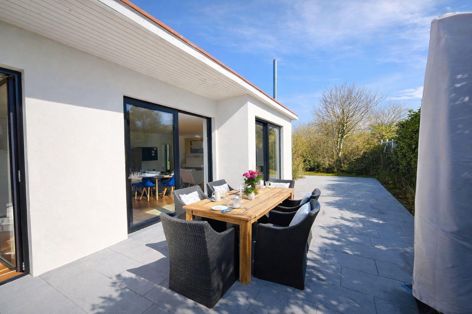 Terrasse mit Holztisch, Stühlen und Blumen. Blick ins Haus durch Glastüren.