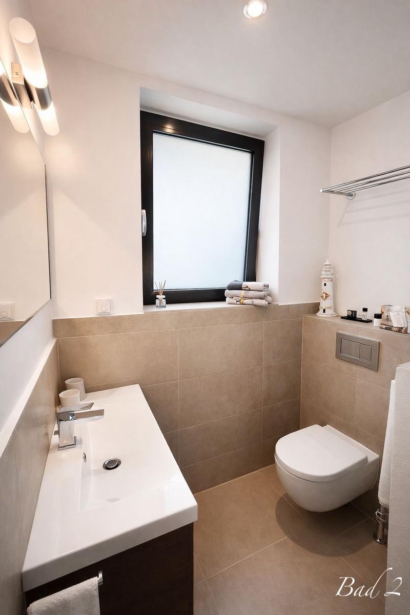 Badezimmer mit Waschbecken, Toilette und Fenster. Fliesenwand und -boden.