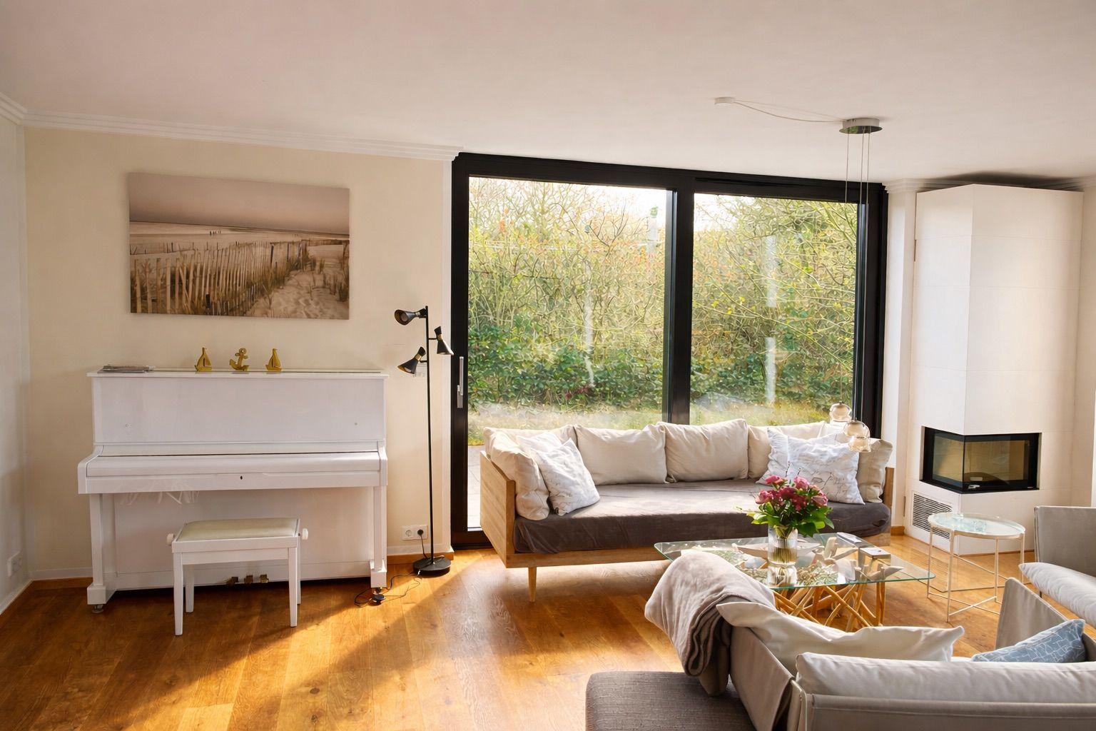 Wohnzimmer mit Klavier, Sofa und Glas-Tisch. Große Fensterfront mit Blick auf Garten.