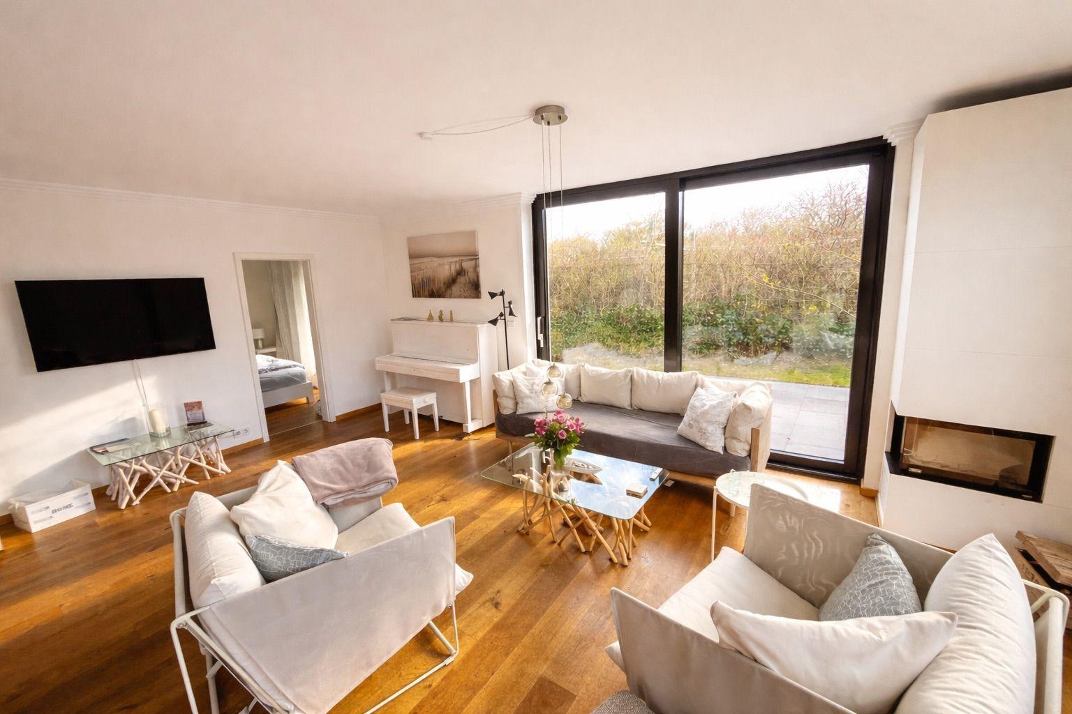Wohnzimmer mit Sofa, Sesseln, Klavier und großem Fenster. Holzboden und Kamin.