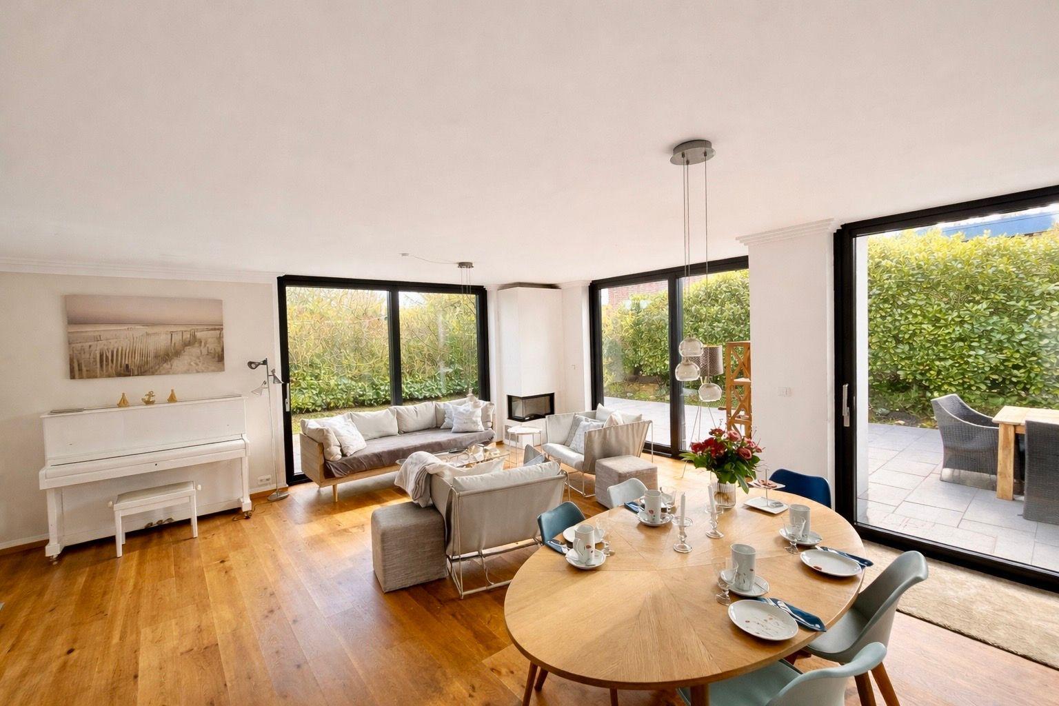 Wohn- und Essbereich mit Piano, Sofa und runden Tisch. Durchgänge zu Terrasse und Garten.