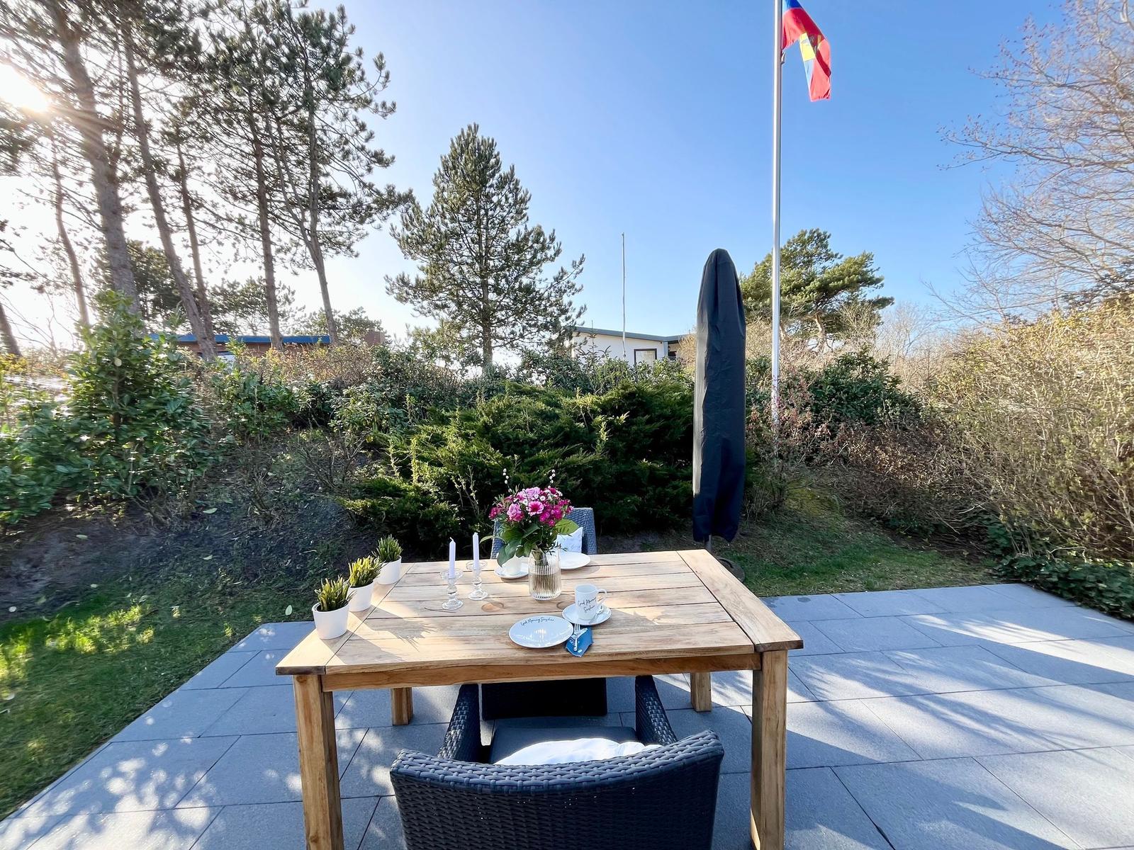 Holztisch mit Stühlen auf Terrasse, Blumen, Flagge und Bäume im Hintergrund.
