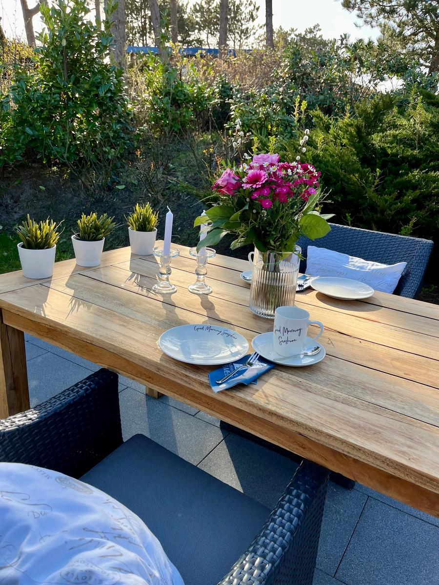 Holztisch mit Blumen, Tassen und Stühlen im Garten.