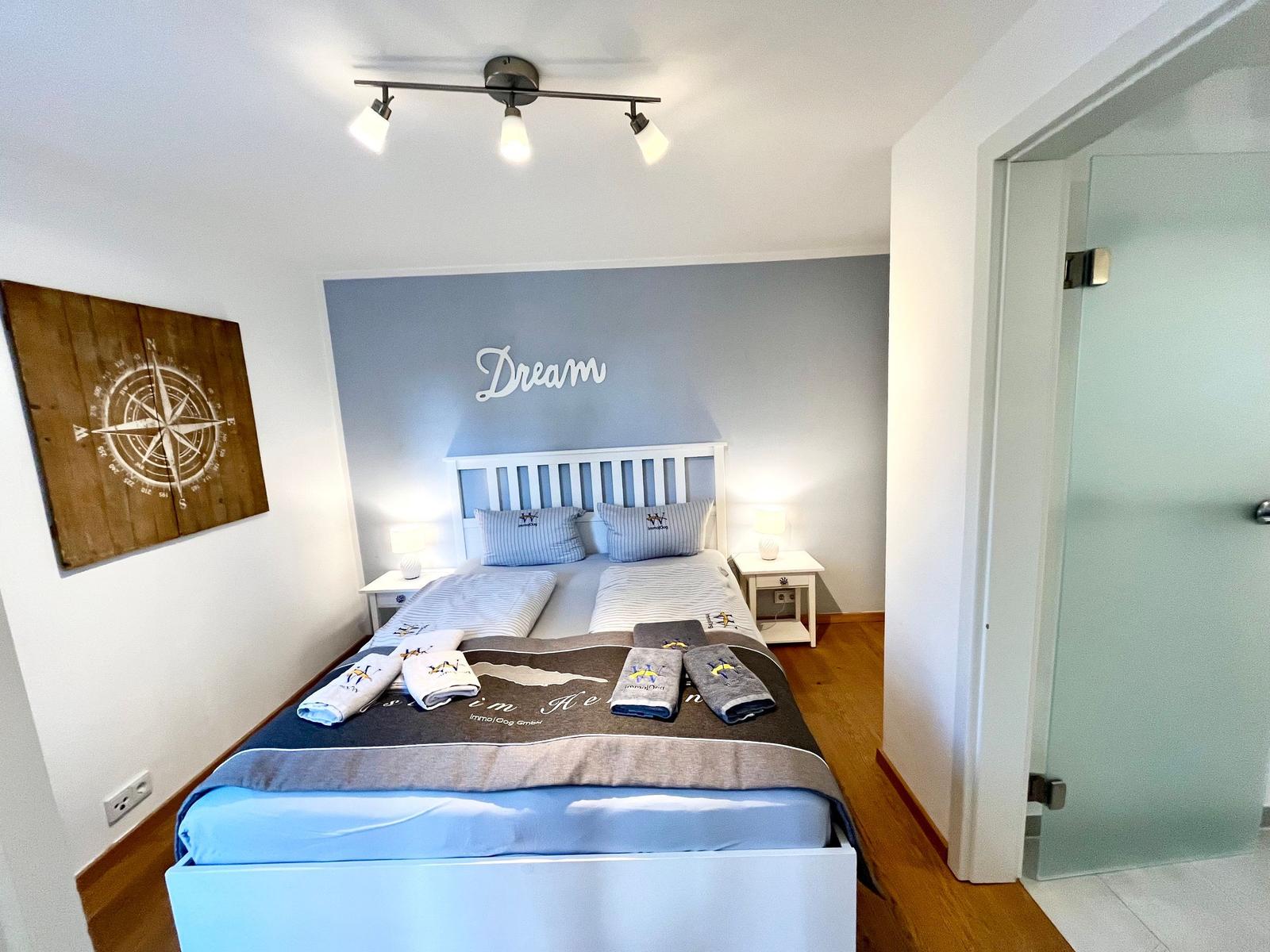 Schlafzimmer mit Bett, Nachttischen und Wanddekoration. Tür führt in Bad.