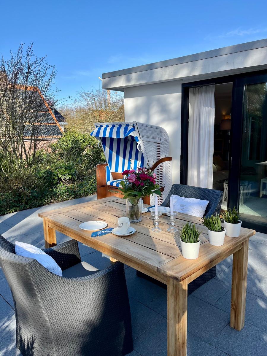 Terrasse mit Holztisch, Stühlen und Strandkorb. Blumen und Pflanzen schmücken den Außenbereich.