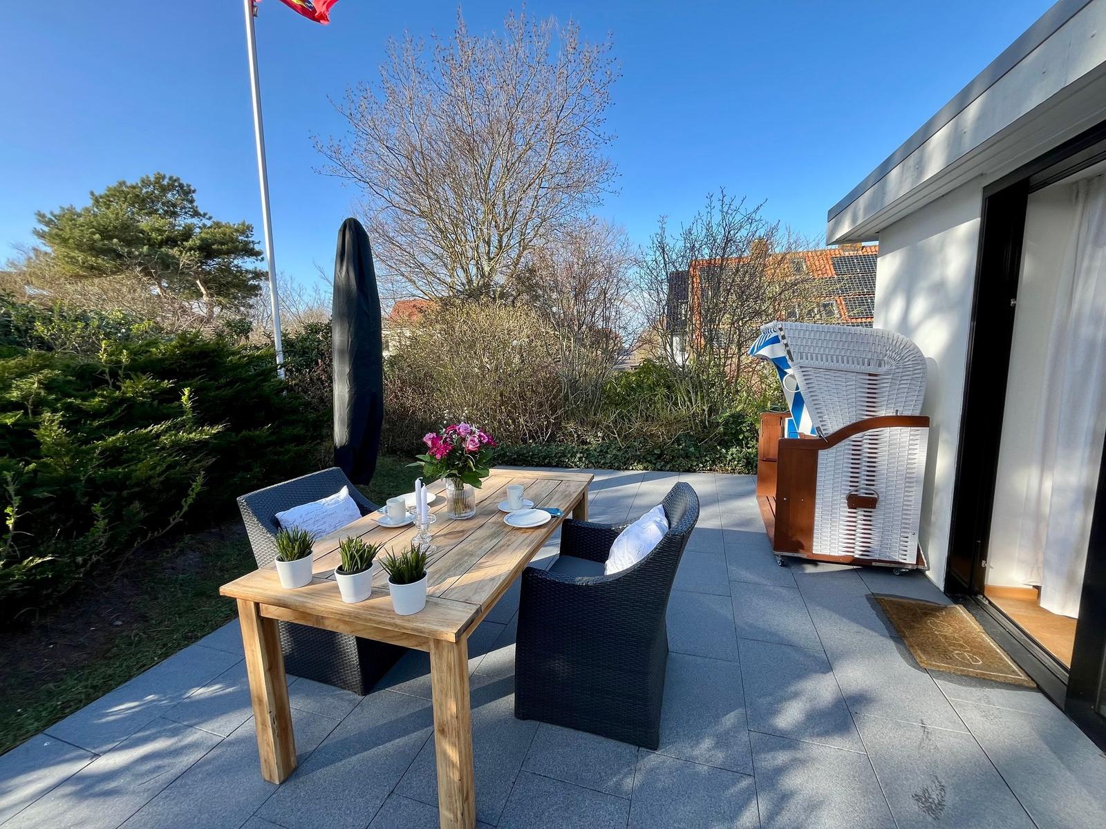 Terrasse mit Tisch, Stühlen, Blumen und Sonnenschirm. Hintergrund mit Bäumen und Haus.